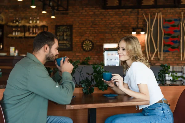Cheerful blonde woman looking at boyfriend drinking coffee during date in cafe — Stock Photo