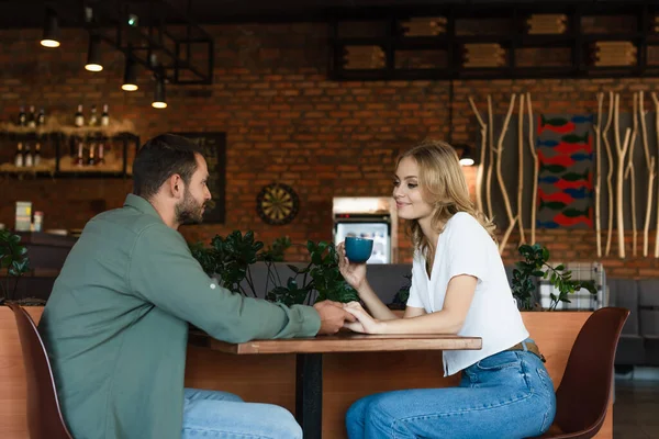 Happy woman with cup of coffee holding hands with boyfriend in cafe — Stock Photo