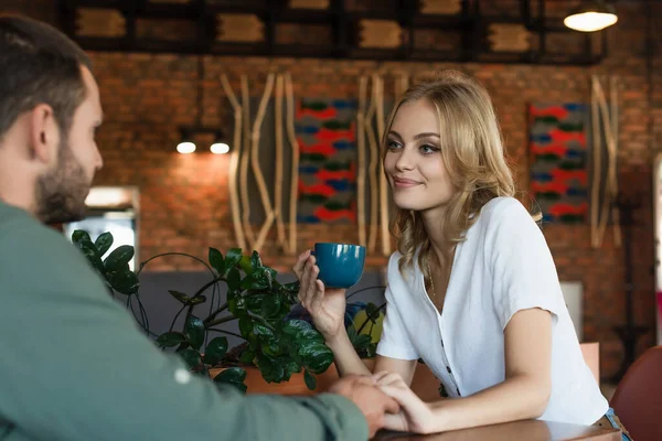 Mujer rubia complacida con taza de café cogido de la mano con novio borroso en la cafetería - foto de stock