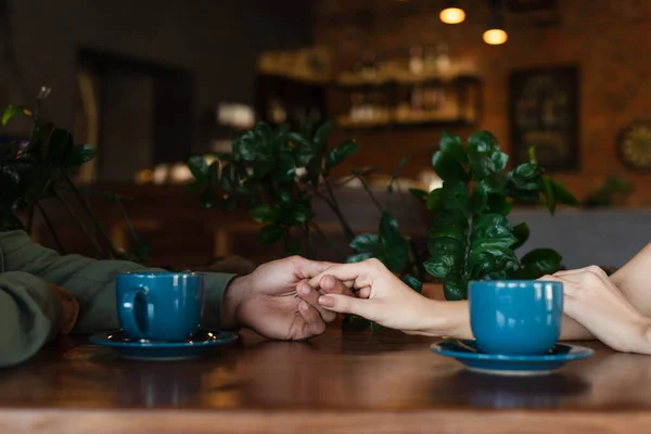 Vista parcial de pareja cogida de la mano cerca de tazas de café durante la cita romántica en la cafetería - foto de stock