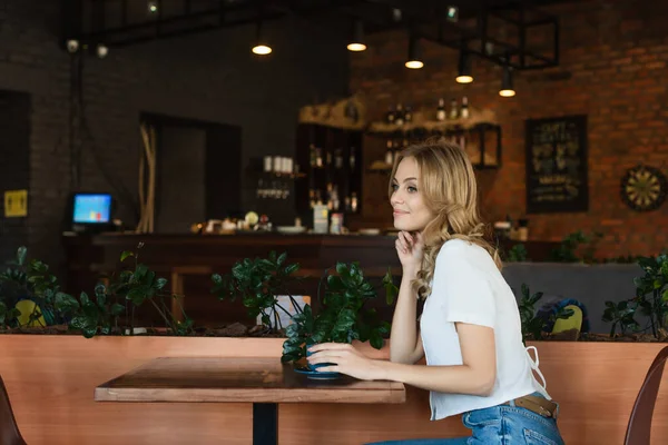 Mujer bonita y feliz mirando hacia otro lado mientras está sentado en la cafetería cerca de la taza de café - foto de stock