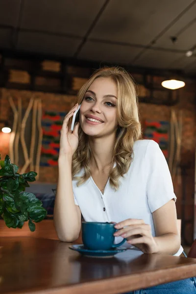 Mujer rubia alegre hablando en el teléfono móvil cerca de la taza de café - foto de stock