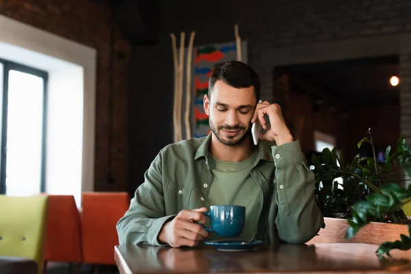Улыбающийся мужчина держит чашку кофе, разговаривая по сотовому телефону в кафе — стоковое фото