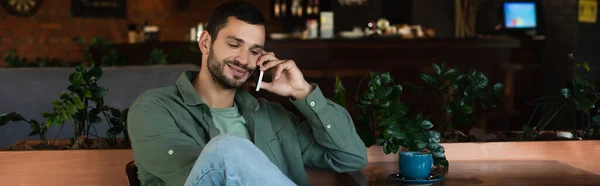 Positive young man talking on cellphone while sitting in restaurant, banner — Stockfoto