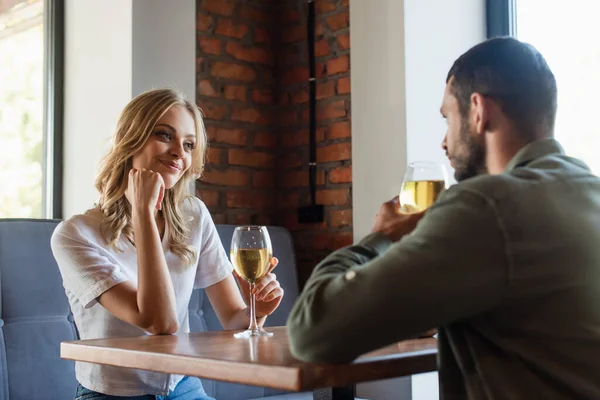 Mujer alegre mirando al hombre borroso mientras bebe vino blanco en el restaurante - foto de stock