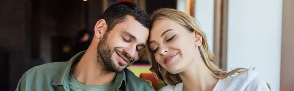 Joven pareja sonriendo con los ojos cerrados mientras se sienta cabeza a cabeza en la cafetería, pancarta - foto de stock