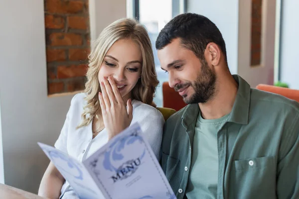 Fröhliche Frau, die Mund mit der Hand bedeckt, während sie mit ihrem Freund Menü liest — Stockfoto