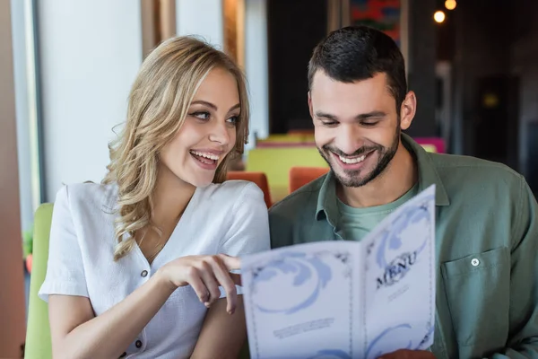 Fröhliche Frau zeigt mit dem Finger auf lächelnden Freund, der Mahlzeit aus Speisekarte auswählt — Stockfoto