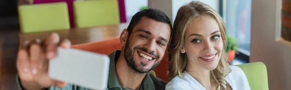 Cheerful man taking selfie on blurred smartphone with happy blonde woman, banner — Stock Photo