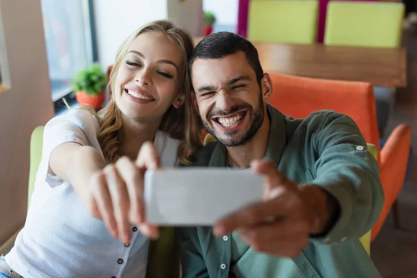 Homem animado sorrindo perto de mulher alegre ao tomar selfie no smartphone borrado — Fotografia de Stock