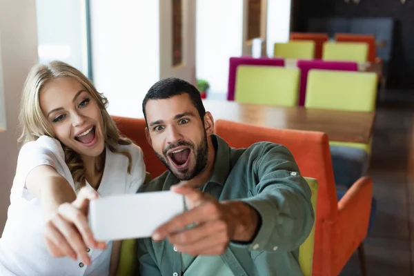 Pareja emocionada divirtiéndose mientras toma selfie en el teléfono celular borroso en la cafetería - foto de stock