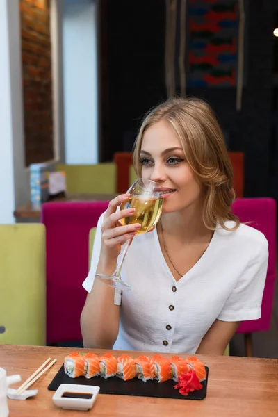 Jovem e feliz mulher bebendo vinho branco perto de saborosos rolos de sushi — Fotografia de Stock