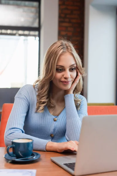 Pleased woman using laptop near coffee cup in restaurant — Foto stock