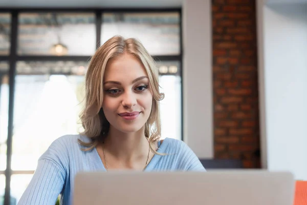 Alegre mujer rubia mirando borrosa portátil en la cafetería - foto de stock