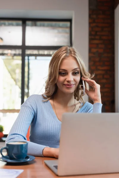 Happy woman talking on mobile phone near blurred laptop and cup of coffee — Fotografia de Stock