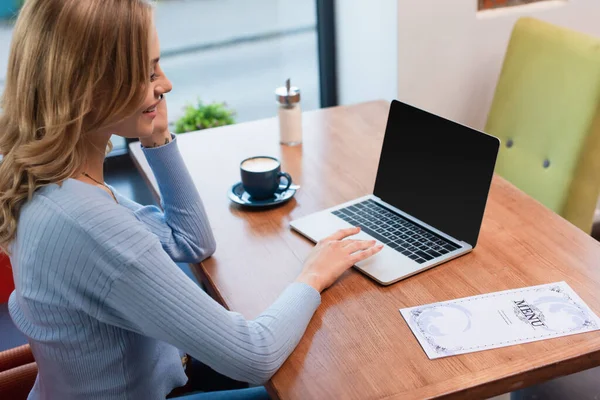 Donna felice seduta vicino al computer portatile con schermo bianco, tazza di caffè e menu sul tavolo — Foto stock