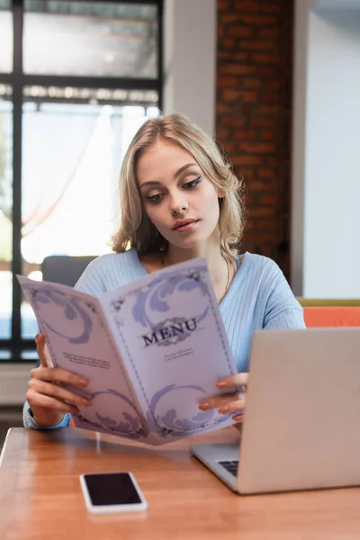 Young blonde woman reading menu near laptop and smartphone with blank screen — Stock Photo