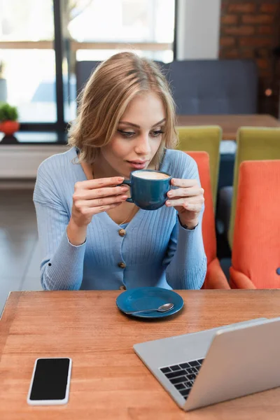 Mulher bonita bebendo café enquanto olha para laptop perto de celular com tela em branco — Fotografia de Stock