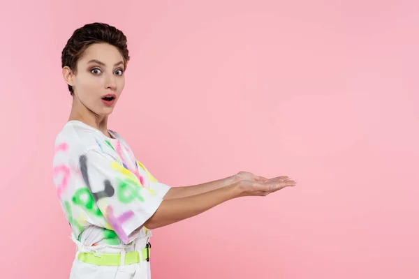 Astonished woman in stylish t-shirt looking at camera and pointing with hands isolated on pink — Stockfoto