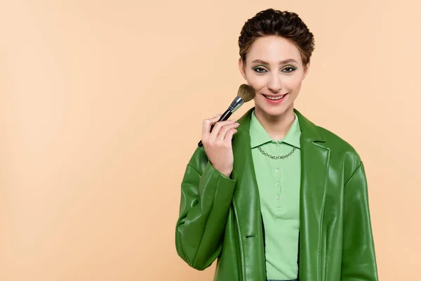 Femme heureuse en veste verte appliquant de la poudre visage avec pinceau cosmétique isolé sur beige — Photo de stock