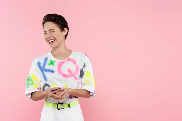 Mujer excitada con los ojos cerrados riendo mientras sostiene el teléfono inteligente aislado en rosa - foto de stock