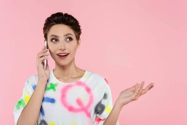 Mujer feliz con el pelo corto apuntando con la mano mientras habla en el teléfono móvil aislado en rosa - foto de stock