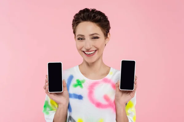 Young brunette woman showing mobile phones while smiling at camera isolated on pink — Fotografia de Stock