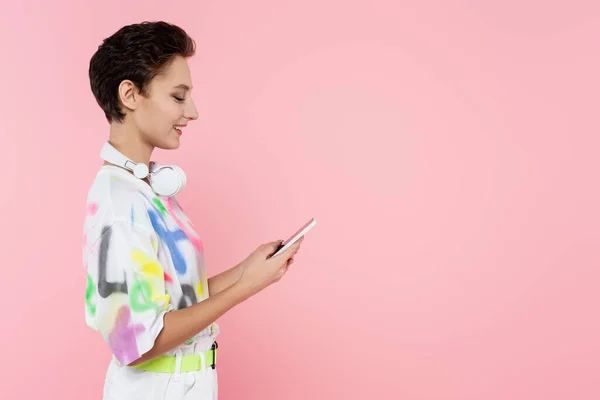 Seitenansicht einer lächelnden Frau mit drahtlosen Kopfhörern am Hals, die ihr Handy isoliert auf rosa verwendet — Stockfoto