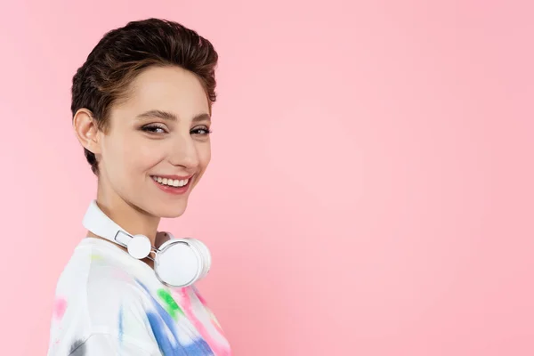 Cheerful woman with wireless headphones on neck looking at camera isolated on pink — стоковое фото