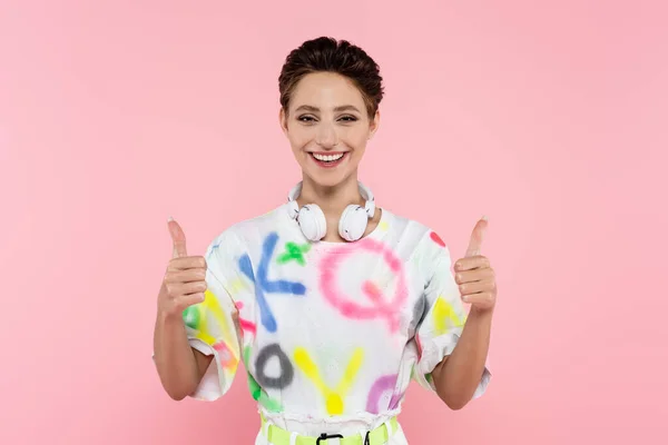 Happy woman with headphones showing thumbs up while looking at camera isolated on pink — Stockfoto