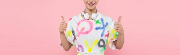 Vista recortada de la mujer feliz con auriculares en el cuello mostrando los pulgares hacia arriba aislado en rosa, bandera - foto de stock
