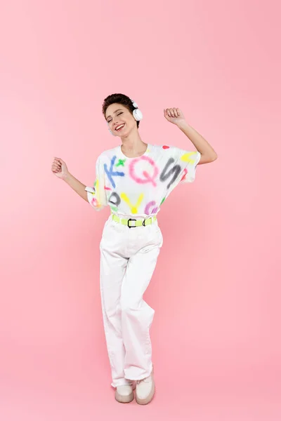 Vista completa de la mujer con estilo feliz con los ojos cerrados bailando en auriculares en rosa - foto de stock