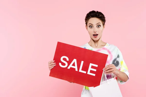 Begeisterte Frau mit Einkaufstasche und roter Karte mit Verkaufsaufdruck, die isoliert in die Kamera auf rosa schaut — Stockfoto