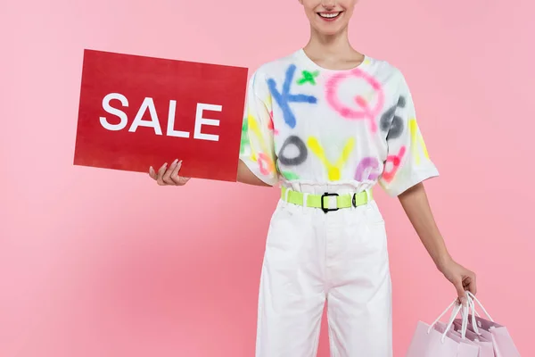 Vista parcial de una mujer sonriente sosteniendo bolsas de compras y tarjeta roja con letras de venta aisladas en rosa - foto de stock