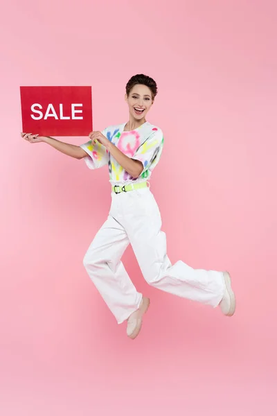 Excited woman in stylish clothes jumping with red card with sale lettering on pink — Foto stock