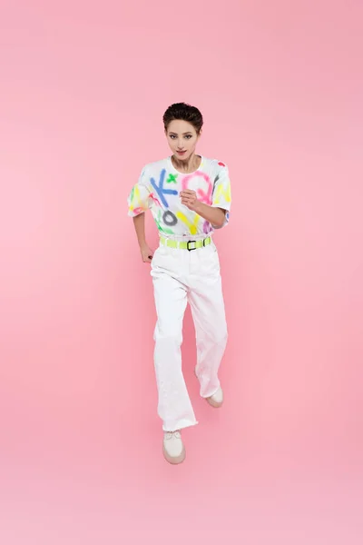 Vista completa de la mujer en camiseta de moda y pantalones blancos corriendo en rosa - foto de stock