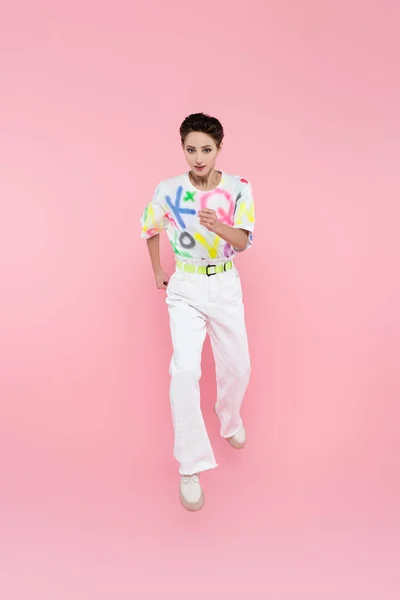 Vista completa de la mujer en camiseta de moda y pantalones blancos corriendo en rosa — Stock Photo