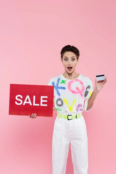 Verblüffte Frau mit Kreditkarte und rotem Plakat mit Verkaufsaufdruck auf rosa — Stockfoto