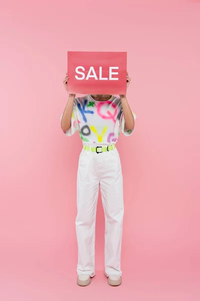 Ganzkörperansicht einer Frau in weißer Hose, die ihr Gesicht hinter einer Karte mit Verkaufsaufdruck auf rosa verdeckt — Stockfoto