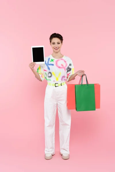 Vista completa de la mujer feliz sosteniendo bolsas de compras y tableta digital con pantalla en blanco en rosa - foto de stock