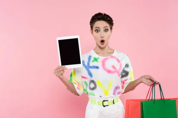 Mujer morena asombrada sosteniendo bolsas de compras y tableta digital con pantalla en blanco aislada en rosa - foto de stock