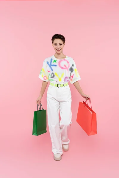 Вид в полный рост счастливой женщины в белых брюках, идущей с сумками для покупок на розовом фоне — стоковое фото