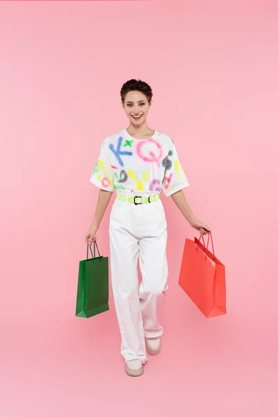 Full length view of happy woman in white trousers walking with shopping bags on pink — Foto stock