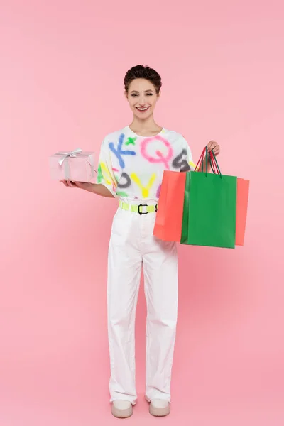 Повний вигляд стильної та радісної жінки, яка позує з подарунковою коробкою та сумками для покупок на рожевому — стокове фото