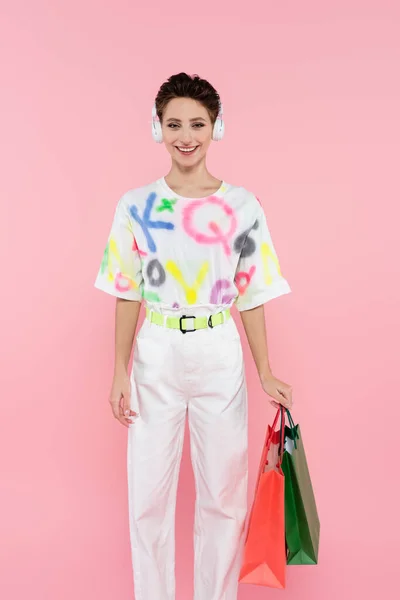 Young and cheerful woman in headphones standing with shopping bags isolated on pink — Foto stock