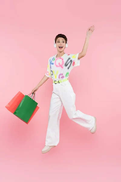 Vista completa de la mujer alegre en auriculares levitando con bolsas de compras mientras agita la mano en rosa - foto de stock