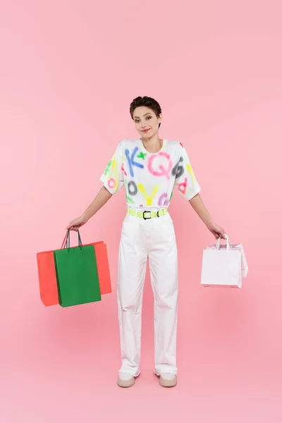 Вид в полный рост улыбающейся женщины с разноцветными сумками для покупок, смотрящей в камеру на розовом фоне — стоковое фото