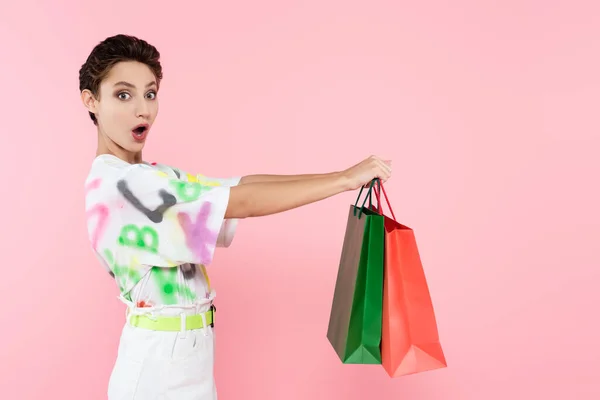 Astonished woman with open mouth holding shopping bags and looking at camera isolated on pink — Stockfoto