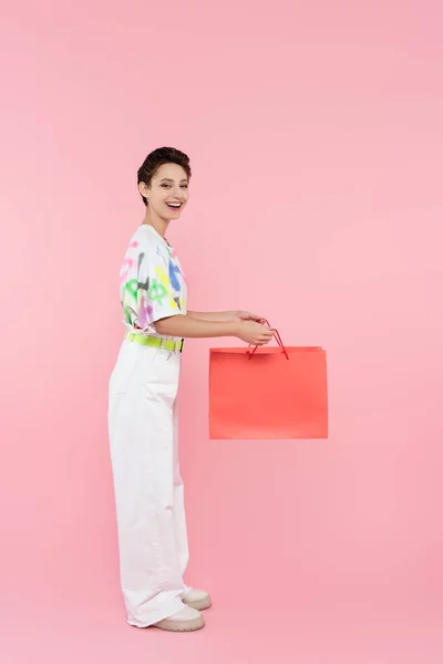 Вид в полный рост счастливой женщины в белых брюках, держащей сумку с покупками на розовом фоне — стоковое фото
