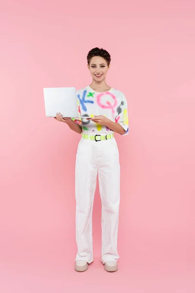 Vue pleine longueur de femme heureuse en pantalon blanc debout avec ordinateur portable sur rose — Photo de stock